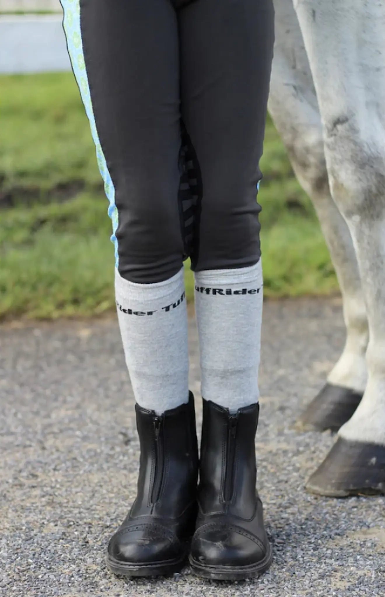 Person wearing black riding boots and gray socks with 'TuffRider' branding, standing next to a horse.